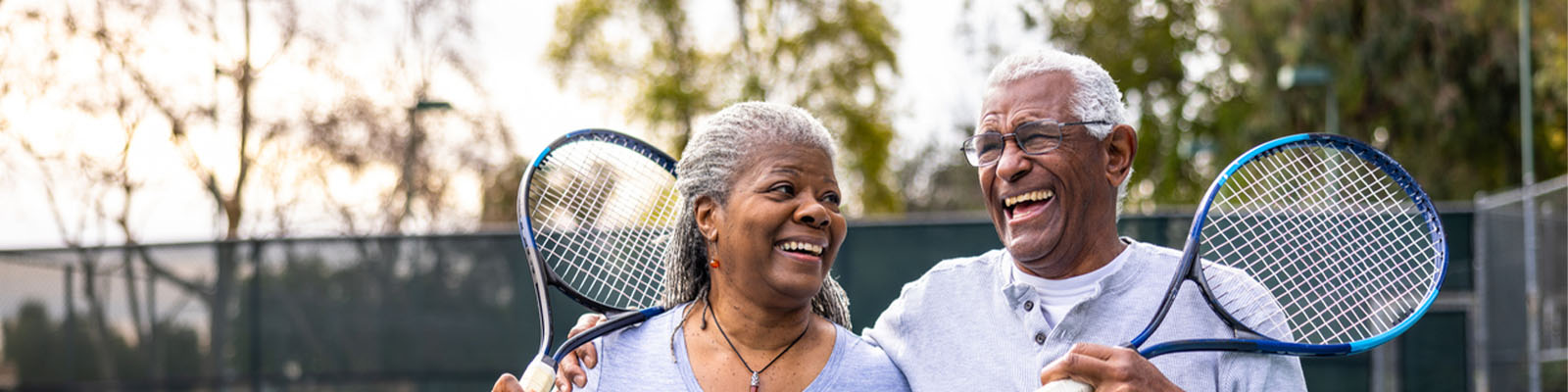 An elderly senior living couple standing on a tennis court holding tennis rackets while smiling
