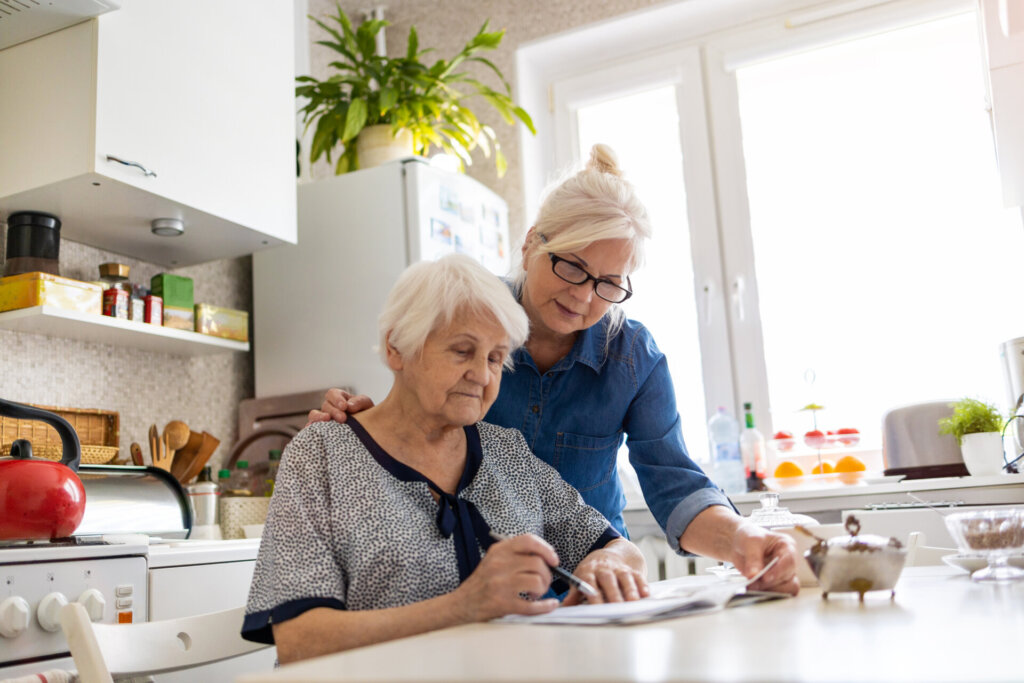 A mature woman assisting her elderly mother with paperwork at a kitchen table