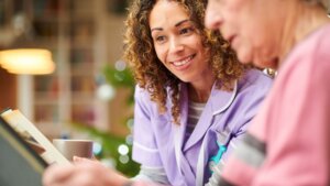 A senior woman wearing a pink shirt looking down at a photo album while sitting beside her female memory care nurse